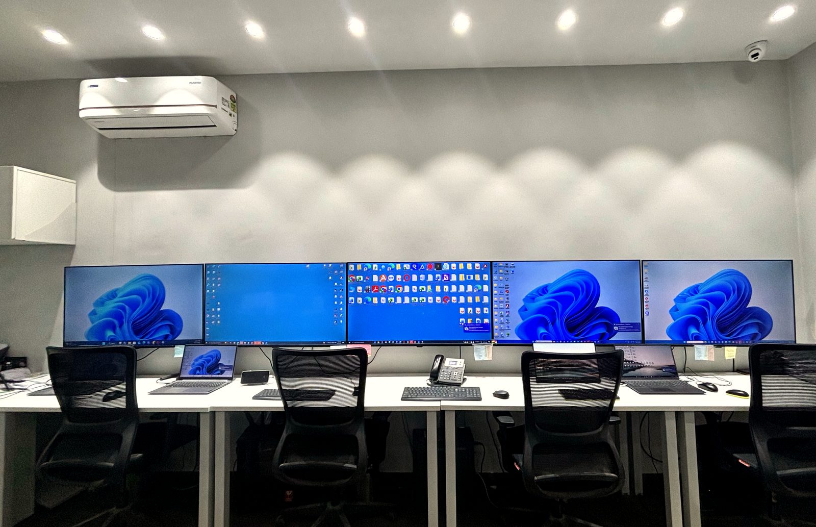 A row of five workstations along a single desk, each with a large monitor, keyboard and office chair, in a lit and air-conditioned room with a desk phone at the centre station.